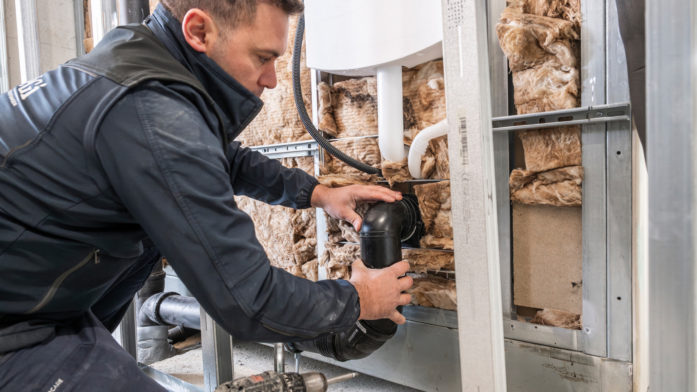 Ein Fachhandwerker montiert in einem geöffneten Wandbereich die Abwasserleitung eines Badezimmers. Sichtbar sind Metallständerwerk, Dämmung, Rohre und ein Bohrschrauber.